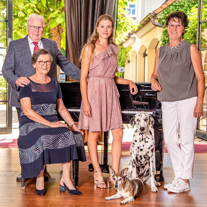 Hotel Weisses Lamm Veitsh&ouml;chheim - Familie Prescher mit Tochter Anna, Heike Prescher und Eltern, sowie zwei Hunde vor einem Klavierfl&uuml;gel in einem hellen Raum mit gro&szlig;en Fenstern und gr&uuml;nen Pflanzen im Hintergrund
