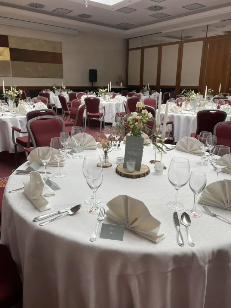 Festlich gedeckte runde Tische mit weißen Tischdecken, gefalteten Servietten, Gläsern, Besteck und Blumenarrangements in einem Bankettsaal mit roten Stühlen