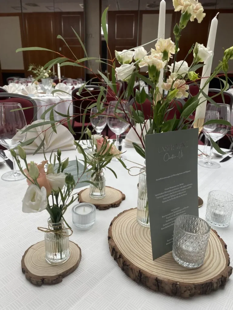 Tischdekoration mit Holzscheiben, Glasvasen mit weißen und rosa Blumen, Kerzen und einem grauen Menü auf einem festlich gedeckten Tisch
