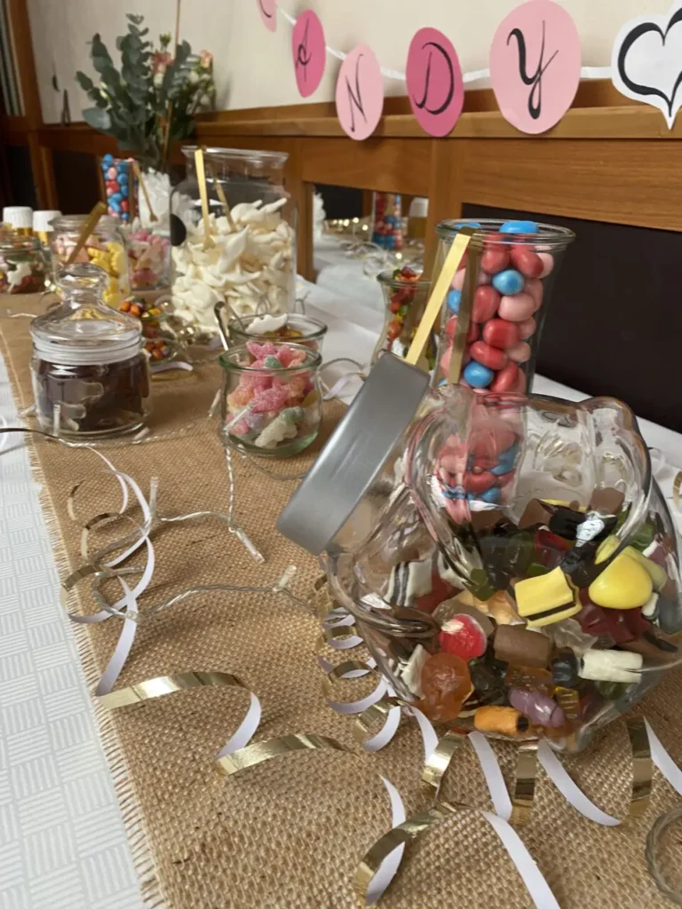 Tisch mit verschiedenen Gläsern und Schalen voller bunter Süßigkeiten und dekorativen Bändern auf einer Jutetischdecke vor einer Holzbank mit rosa Buchstaben-Dekoration
