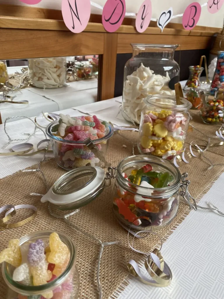 Mehrere Gläser mit verschiedenen bunten Süßigkeiten auf einem Tisch mit Jutedecke und silbernen Luftschlangen
