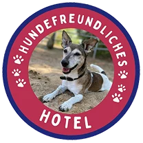 Rundes rotes Abzeichen mit wei&szlig;er Schrift 'Hundefreundliches Hotel' und sechs Pfotenabdr&uuml;cken, im Zentrum ein liegender Hund mit braun-wei&szlig;em Fell und schwarzer Schnauze.